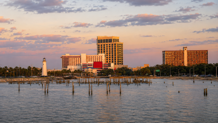 Biloxi Beach with the historic lighthouse, Gulf Coast shoreline, and casino resorts in the distance on a sunny day.