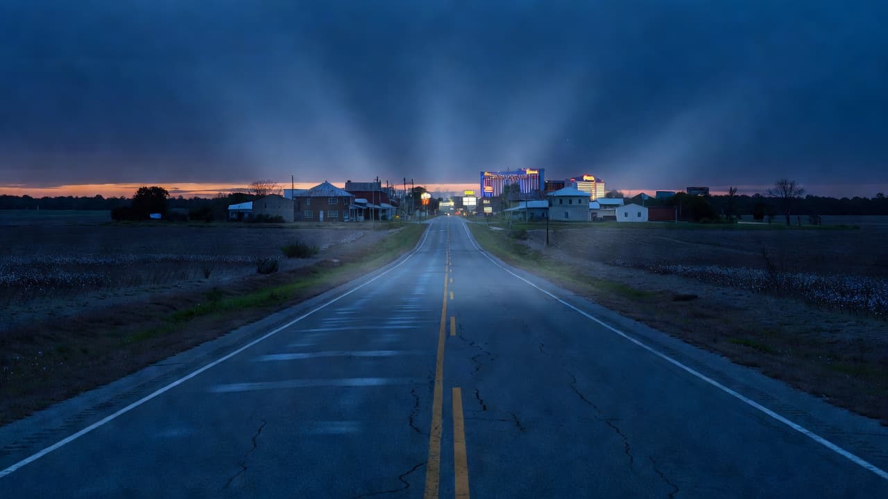 An empty asphalt road stretches toward a distant Mississippi town under deep blue twilight skies. Low-lying fields flank the desolate highway, creating a cinematic and moody atmosphere with stark architectural silhouettes.