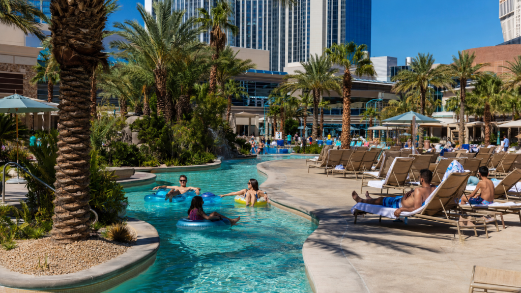 Family-friendly Las Vegas resort pool complex with lounge chairs, palm trees, lazy river-style water feature, and resort towers.